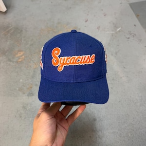 Op de afbeelding: Een koningsblauwe baseballpet met het woord Syracuse in oranje schrijfletters met een witte omlijning op het voorpaneel geborduurd. De pet heeft extra geborduurde patches op de zijkanten en een gestructureerde kroon met een gebogen klep.