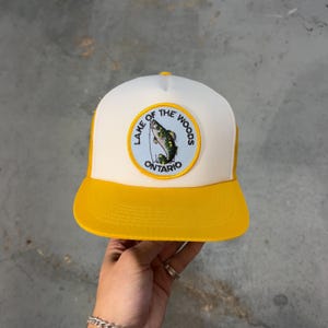 Peut inclure: Casquette de camionneur jaune et blanche avec une visière incurvée. Le chapeau présente un écusson circulaire avec un graphique de poisson et le texte "Lake of the Woods Ontario". Le chapeau a un dos en filet.