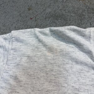 Puede incluir: Primer plano de una camiseta gris claro con un patr&oacute;n moteado. La tela tiene una textura suave y muestra una ligera decoloraci&oacute;n. Se ven el cuello y la costura del hombro.