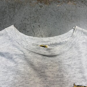 Puede incluir: Una camiseta gris jaspeada con cuello redondo. La camiseta tiene una etiqueta en el cuello. La tela tiene un aspecto moteado. La camiseta es probablemente vintage o de segunda mano.