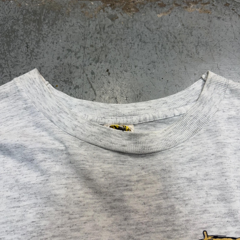 Puede incluir: Una camiseta gris jaspeada con cuello redondo. La camiseta tiene una etiqueta en el cuello. La tela tiene un aspecto moteado. La camiseta es probablemente vintage o de segunda mano.