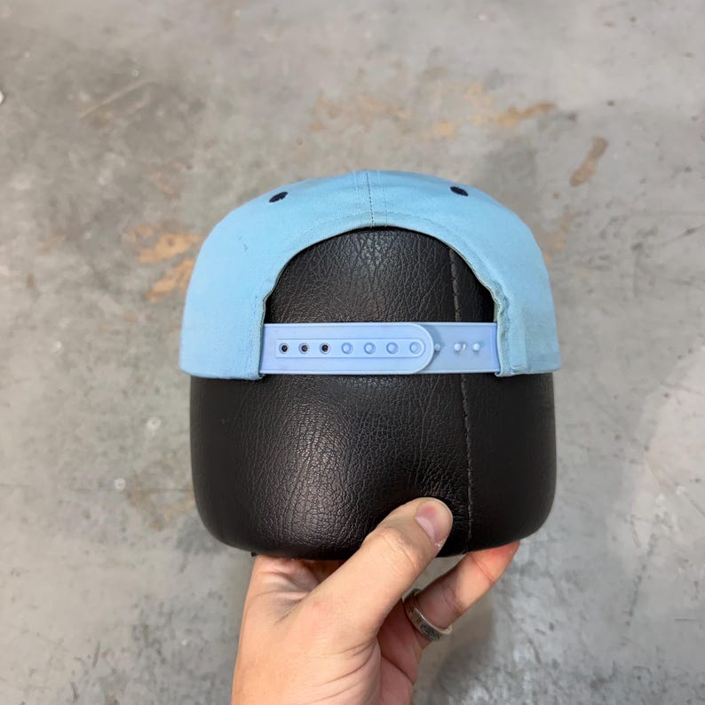 May include: A light blue baseball cap with a curved brim and adjustable strap. The cap is displayed from the back, showcasing the strap's adjustment holes and the cap's solid color design. The cap is on a black surface.