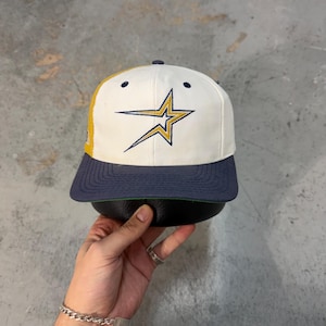Puede incluir: Gorra de béisbol con un panel frontal blanco con un logotipo de estrella dorada y azul marino. La visera es azul marino y la parte trasera de malla es amarilla. La gorra tiene un cierre a presión.