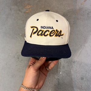 May include: Cream-colored Indiana Pacers baseball cap with a navy blue brim. The team name "Pacers" is in gold script with a navy blue outline. The word "Indiana" is above the team name.