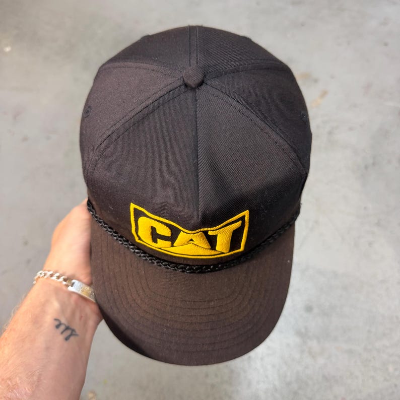 Może przedstawiać: Czarna czapka bejsbol&oacute;wka z ż&oacute;łto-czarnym logo CAT. Czapka ma zakrzywiony daszek, sznurkowy detal i guzik na g&oacute;rze. Klasyczna czapka w stylu trucker.