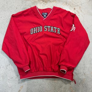 Könnte beinhalten: Roter Ohio State Buckeyes Windbreaker mit weißem Besatz. Die Worte "OHIO STATE" stehen in weißen Blockbuchstaben auf der Brust. Das Ohio State-Logo befindet sich auf dem rechten Ärmel. Der Windbreaker hat einen V-Ausschnitt und elastische Bündchen.
