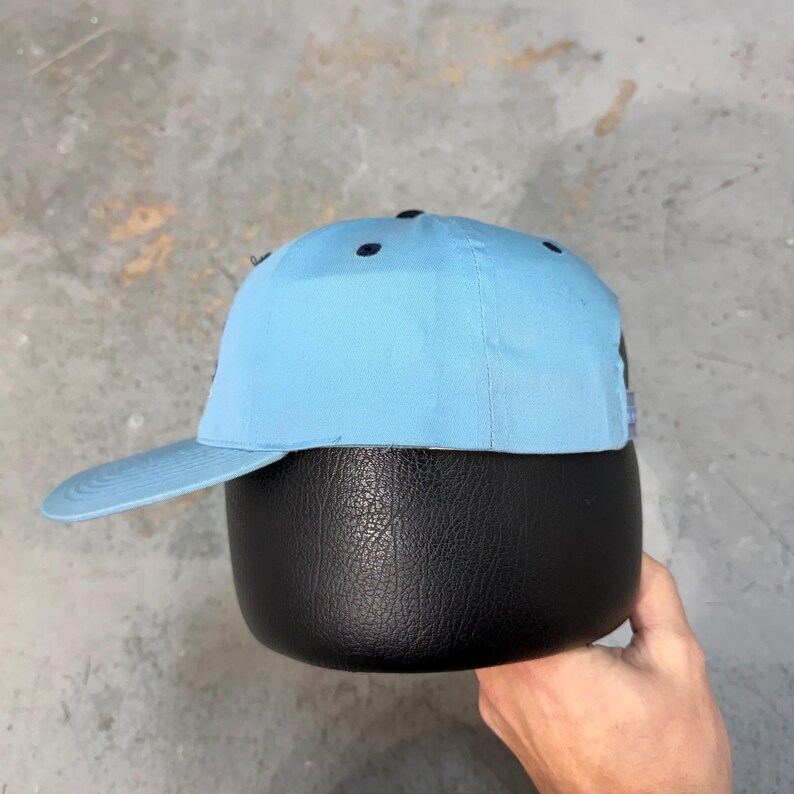 May include: A light blue baseball cap with a curved brim. The cap has a structured crown with six ventilation eyelets. The cap is displayed on a black, rounded object. The background is a mottled gray surface.