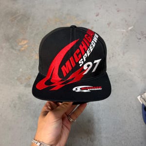 Può includere: Un cappellino da baseball nero con scritta ricamata in rosso e bianco che recita Michigan Speedway e il numero 97. Il cappellino presenta una visiera piatta e un logo Michigan Speedway più piccolo in bianco e rosso sulla parte anteriore.