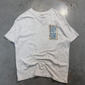 Puede incluir: Camiseta gris jaspeado con el texto "BODY GLOVE" en un dise&ntilde;o rectangular turquesa y dorado en el pecho. La camiseta tiene mangas cortas y cuello redondo. La tela parece suave y ligeramente desgastada.