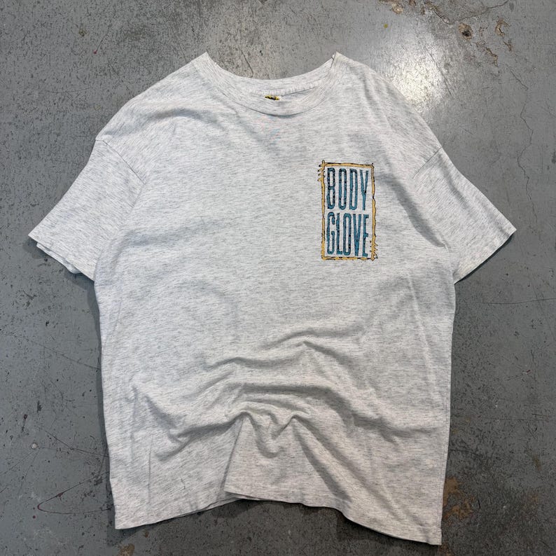 Puede incluir: Camiseta gris jaspeado con el texto "BODY GLOVE" en un dise&ntilde;o rectangular turquesa y dorado en el pecho. La camiseta tiene mangas cortas y cuello redondo. La tela parece suave y ligeramente desgastada.