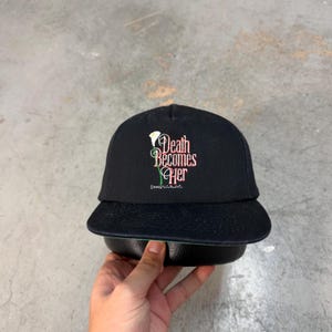 May include: Black baseball cap with a flat brim. The cap features the phrase "Death Becomes Her" in a stylized font, with a floral graphic. The cap has a green underside.