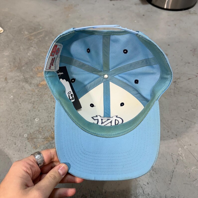 May include: Light blue baseball cap with a flat brim. The interior is light blue and white, with a logo and the letters "LS" on a tag. The cap has a snapback closure and a curved brim.