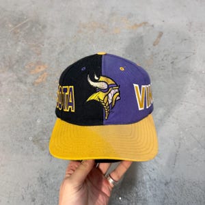 May include: A Minnesota Vikings snapback hat. The hat is split into black and purple panels, with a yellow brim. The team logo and the word "MINNESOTA" are embroidered in yellow. The word "VIKINGS" is embroidered in white.