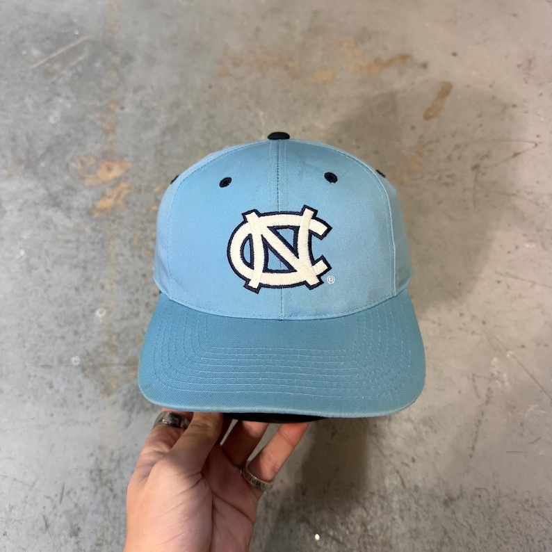 May include: Light blue baseball cap with a curved brim and a black button on top. The cap features a white and navy blue logo on the front. The hat is a classic style, suitable for sports or casual wear.