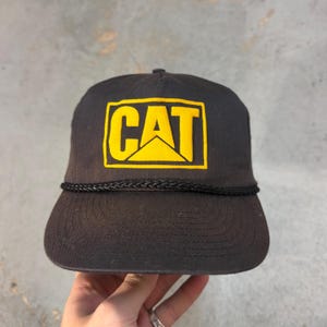 Może przedstawiać: Czarna czapka bejsbolowa CAT z ż&oacute;łtym logo. Czapka ma płaski daszek, czarny sznurkowy detal i klasyczny design czapki typu trucker. Logo CAT znajduje się w ż&oacute;łtym prostokącie.