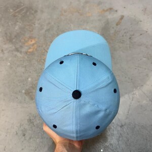 May include: A light blue baseball cap with a curved brim. The cap has a six-panel design with a dark blue button on top and dark blue eyelets. The brim is a solid light blue color.