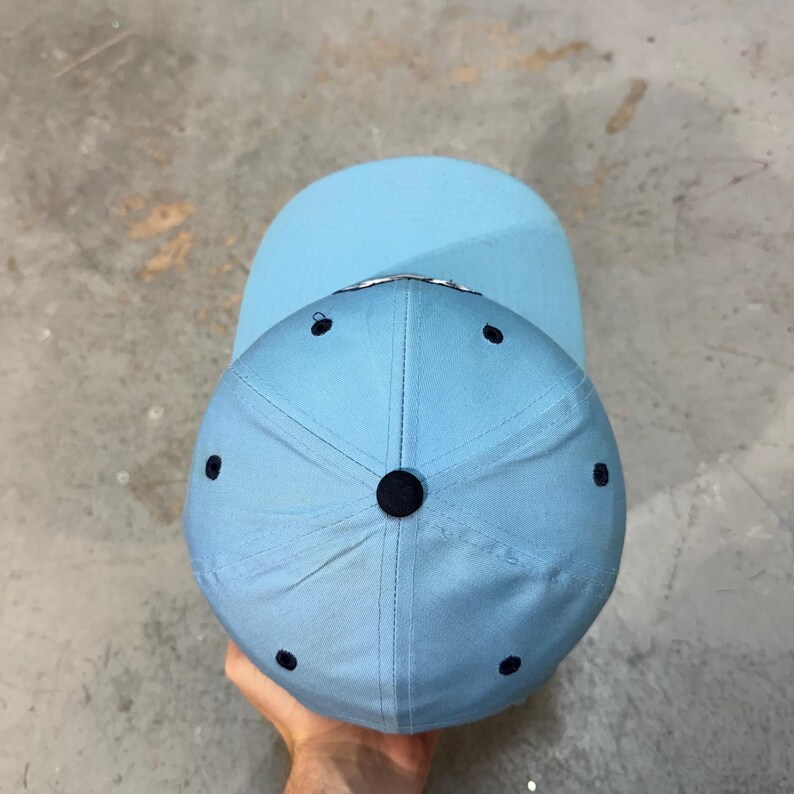 May include: A light blue baseball cap with a curved brim. The cap has a six-panel design with a dark blue button on top and dark blue eyelets. The brim is a solid light blue color.