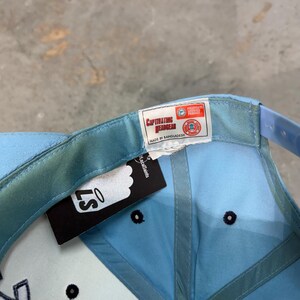 May include: Light blue baseball cap with a curved brim and adjustable strap. The cap features a tag that reads "Captivating Headgear" and "Made in Bangladesh". The cap has a black button on top and a white tag with the text "LS".