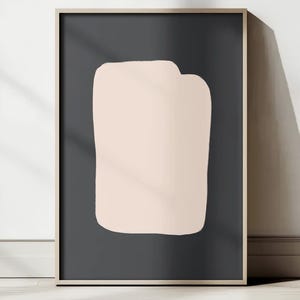 May include: A framed abstract art print featuring a light peach-colored shape against a dark gray background. The artwork is displayed on a white wall and wooden floor, with natural light casting shadows.