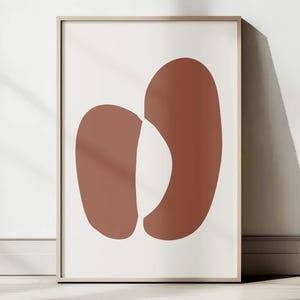 May include: A framed art print featuring two abstract shapes in a warm terracotta color against a white background. The shapes are organic and rounded, with a minimalist design. The frame is a light wood color, and the artwork is displayed on a white wall.