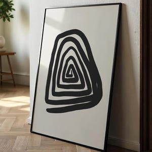 May include: Abstract art print in black and white. The artwork displays a striking black spiral design on a cream background. The print is framed in a thin, black frame and leans against a wall.