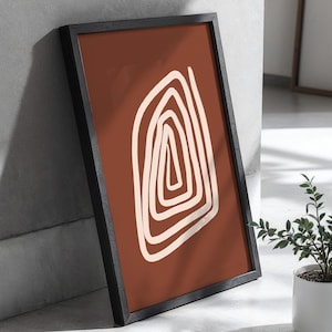 May include: Framed abstract art print featuring a geometric design. The artwork has a terracotta background with a cream-colored, spiral-shaped design. The print is encased in a black frame, leaning against a gray wall.