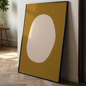 Op de afbeelding: Een ingelijste abstracte kunstprint met een grote, gebroken witte ovale vorm op een mosterdgele achtergrond. Het kunstwerk staat in een zwarte lijst, leunend tegen een witte muur en een houten deurkozijn. De vloer is van hout.