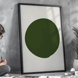 Puede incluir: Un cuadro enmarcado con un gran círculo verde oscuro sobre un fondo blanco. La obra de arte está en un marco negro, apoyada contra una pared de hormigón gris. El diseño minimalista es una pieza de decoración moderna.