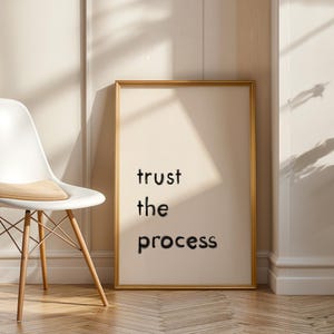 Pode incluir: Uma impressão emoldurada com as palavras "trust the process" em letras pretas. A impressão tem uma moldura dourada e é exibida contra uma parede de cor neutra. Uma cadeira branca com pernas de madeira está em primeiro plano.