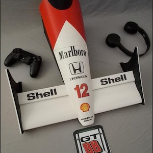 May include: A 3D-printed McLaren Formula 1 car model, with "Marlboro" and "Shell" branding, and the number "12". The model is white, red, and black. A black games controller and black headphones are also visible.