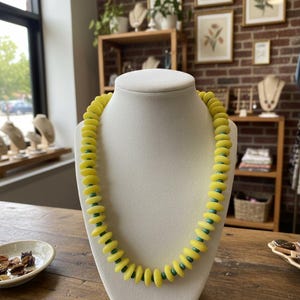 Puede incluir: Un collar de cuentas amarillo con cuentas de acento turquesa exhibido en un maniquí blanco. El collar presenta cuentas circulares planas y grandes. El fondo incluye una mesa de madera, una pared de ladrillo y grabados botánicos enmarcados.