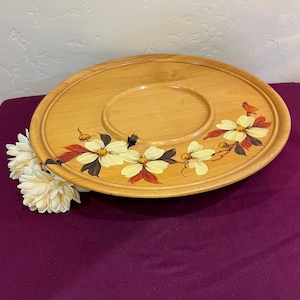 Könnte beinhalten: Ein runder Servierteller aus Holz mit einem zentralen, vertieften Bereich. Der Teller ist mit handgemalten weißen und gelben Blumen mit roten und braunen Akzenten verziert. Ein kleiner Vogel ist auf dem Rand gemalt.