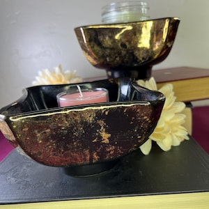 May include: Two black and gold decorative bowls with a candle inside, resting on a stack of books. The bowls have a unique shape and a metallic gold interior. A small pink candle sits in the lower bowl.