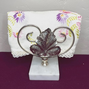 Op de afbeelding: Een decoratieve servethouder met een witte marmeren basis en een zilverkleurig metalen bladontwerp. De houder bevat een wit linnen servet geborduurd met paarse, roze en gele bloempatronen. Het servet heeft een delicate kanten rand.