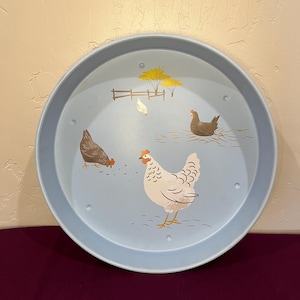 Puede incluir: Una bandeja redonda de color azul claro con una escena de granja pintada. La bandeja presenta ilustraciones de gallinas, una valla y árboles amarillos. Las gallinas son de varios colores, incluyendo blanco, marrón y gris.