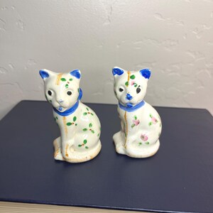 May include: Two vintage ceramic cat ornaments. The white cats have blue ears and collars, with green and pink floral details. They are seated upright on a dark blue surface.