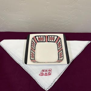 Peut inclure: Un plat carré en céramique blanc cassé avec une bordure noire et un motif géométrique rouge et gris. Il repose sur un mouchoir blanc avec un motif rouge et des coutures noires. Le mouchoir est sur une surface violet foncé.