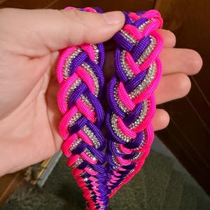 May include: A braided cord accessory featuring vibrant neon pink and deep purple nylon strands interwoven with a shimmering silver rhinestone chain. The design consists of two parallel braided sections that create a textured, multi-colored appearance.