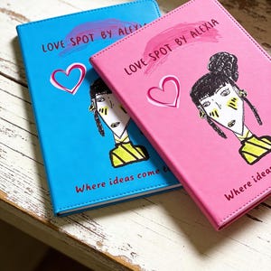 May include: Two notebooks, one blue and one pink, with the text "LOVE SPOT BY ALEXIA" and a heart graphic. Each notebook features a stylized illustration of a woman and the text "Where ideas come to life."