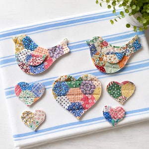 Puede incluir: Una colección de siete adornos de tela acolchada que incluye dos formas de pájaro y cinco formas de corazón. Cada pieza está confeccionada con varios retazos de tela de colores con patrones florales y geométricos en estilo patchwork.