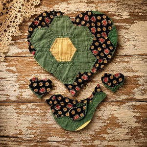 May include: A handmade patchwork heart, two smaller hearts, and a bird-shaped decoration. The heart and bird are green and black with floral patterns. The background is a distressed, weathered wooden surface.