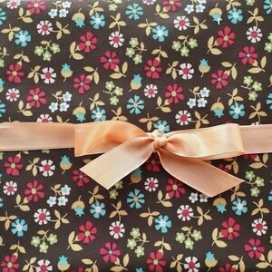 Puede incluir: Una tela con estampado floral sobre un fondo marrón oscuro. El diseño presenta pequeñas flores rojas, blancas, azules y verdes, con hojas amarillas. Una cinta color melocotón está atada en forma de lazo sobre la tela.