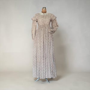 Puede incluir: Un vestido largo de estilo vintage, con cuello alto y mangas largas. El vestido presenta un delicado estampado floral en tonos marrones y blancos. Se exhibe en un maniquí, mostrando su largo total y falda plisada. Las mangas tienen detalles de volantes.