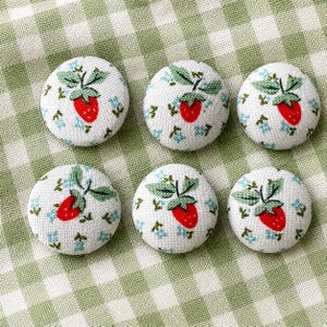 Può includere: Sei bottoni rotondi in tessuto bianco, con un design a fragola rossa con foglie verdi e dettagli floreali blu. I bottoni sono disposti su un tessuto a quadretti verde e bianco.