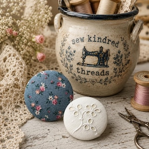 Puede incluir: Una vasija de cerámica con borde y asas azules, con el texto "sew kindred threads" y una ilustración de una máquina de coser. Dos botones forrados de tela: uno azul con estampado floral rosa y otro blanco con bordado de flores.