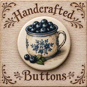 May include: A circular button featuring a printed illustration of a cream-colored ceramic mug filled with dark blue blueberries. The mug has a blue floral pattern and a handle. Two blueberries and green leaves sit at the base of the mug.