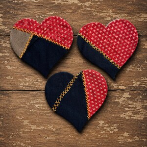 May include: Three heart-shaped fabric ornaments featuring a patchwork design. Each heart is constructed from red fabric with a white cross pattern, solid navy blue fabric, and accents of tan fabric, all joined by visible yellow decorative stitching.