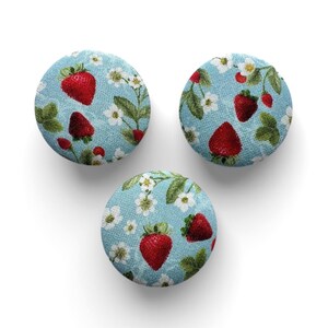 Puede incluir: Tres botones redondos de tela con fondo azul claro. Cada botón presenta un patrón de fresas rojas, flores blancas y hojas verdes. Los botones están dispuestos sobre una superficie blanca.
