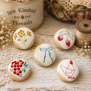 Puede incluir: Cinco botones redondos cubiertos de tela con diseños bordados sobre un fondo color crema. Los motivos incluyen flores amarillas, fresas rojas, un lazo azul, flores rojas con hojas verdes y el contorno rojo de una gallina.