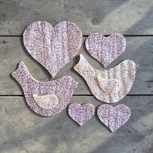 May include: A selection of fabric heart and bird-shaped decorations. The decorations have a lavender floral pattern on a light background. The birds have a yellow underside and a white belly. The decorations are arranged on a weathered wooden surface.